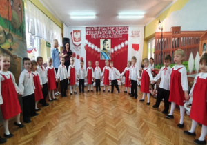 Dziewczynki w białych bluzkach i czerwonych sukienkach oraz chłopcy w białych koszulach i czarnych spodniach w pólkolu śpiewają piosenkę "O mój rozmarynie".